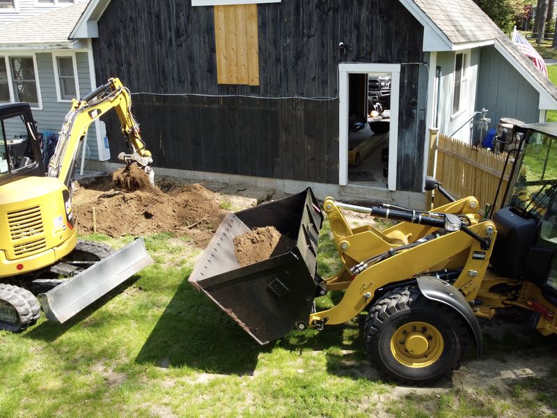 Mini excavator and wheel loader excavating along a barn foundation